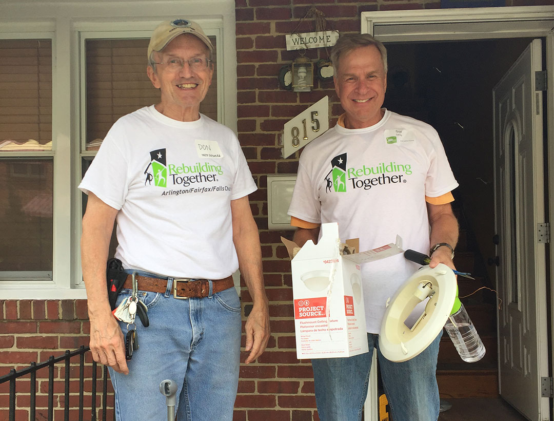 Two volunteers standing on a porch, smiling and holding an empty box of a lighting fixture.